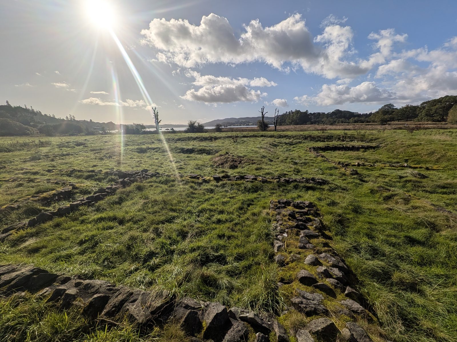 Low stone foundations of Ambleside Roman Fort in rough grass, with Windermere beyond under a bright sun