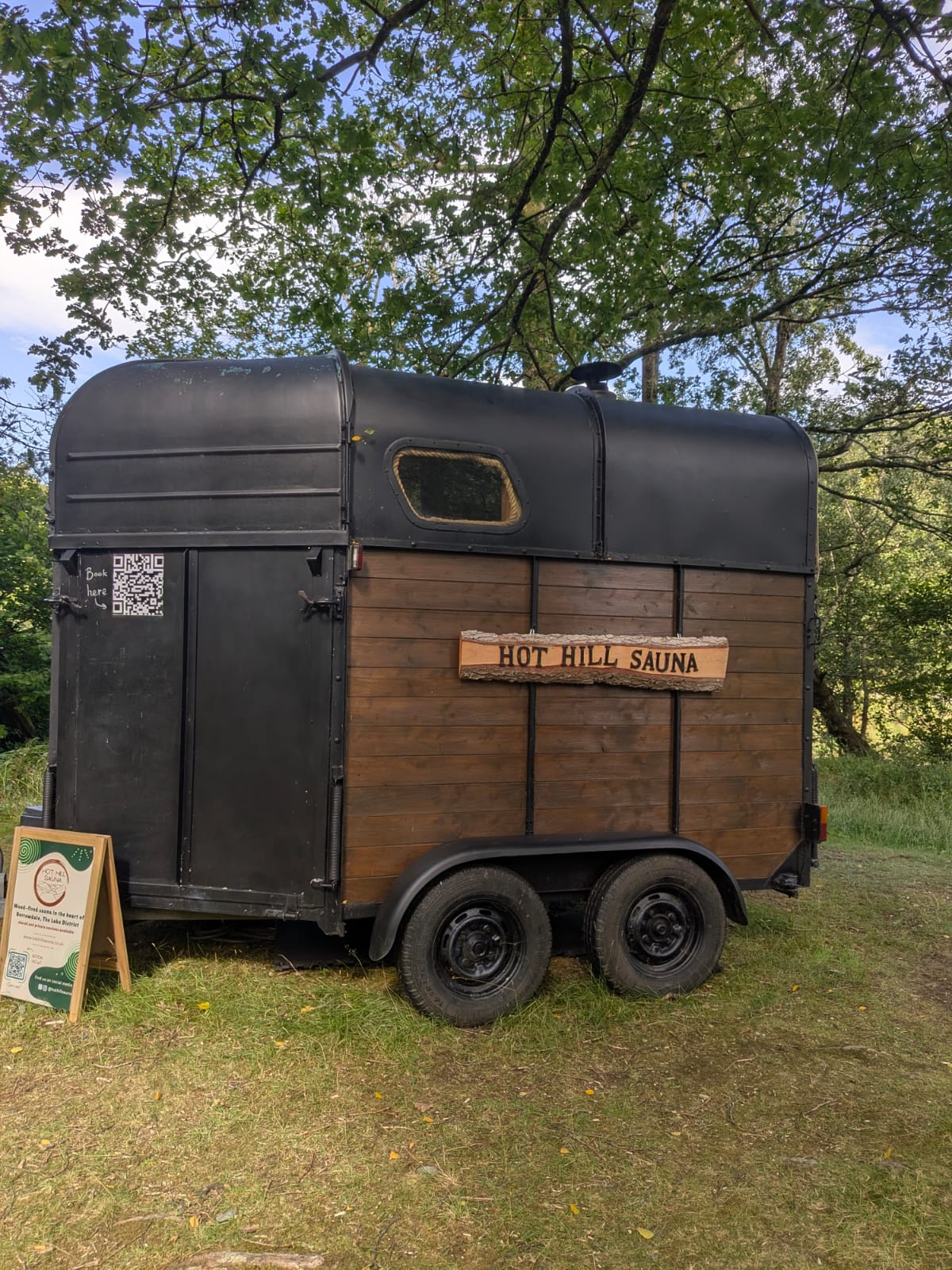 Hot Hill Sauna, a wood-clad converted horsebox sauna parked under trees beside the River Derwent in Borrowdale, with an A-board outside