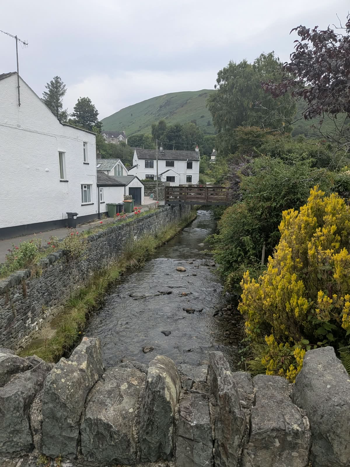 Coledale Beck running between stone walls through Braithwaite village, with a wooden footbridge and whitewashed cottages