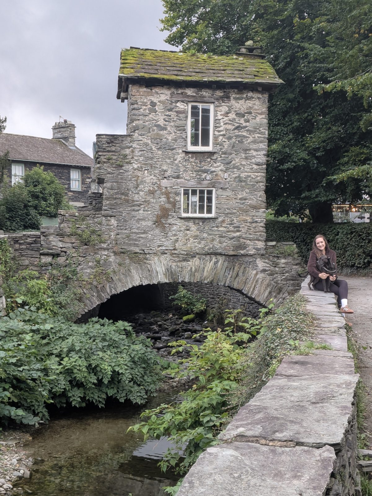 Bridge House over Stock Ghyll, Ambleside, with a person and Cass the Staffordshire Bull Terrier sitting on the stone wall beside it