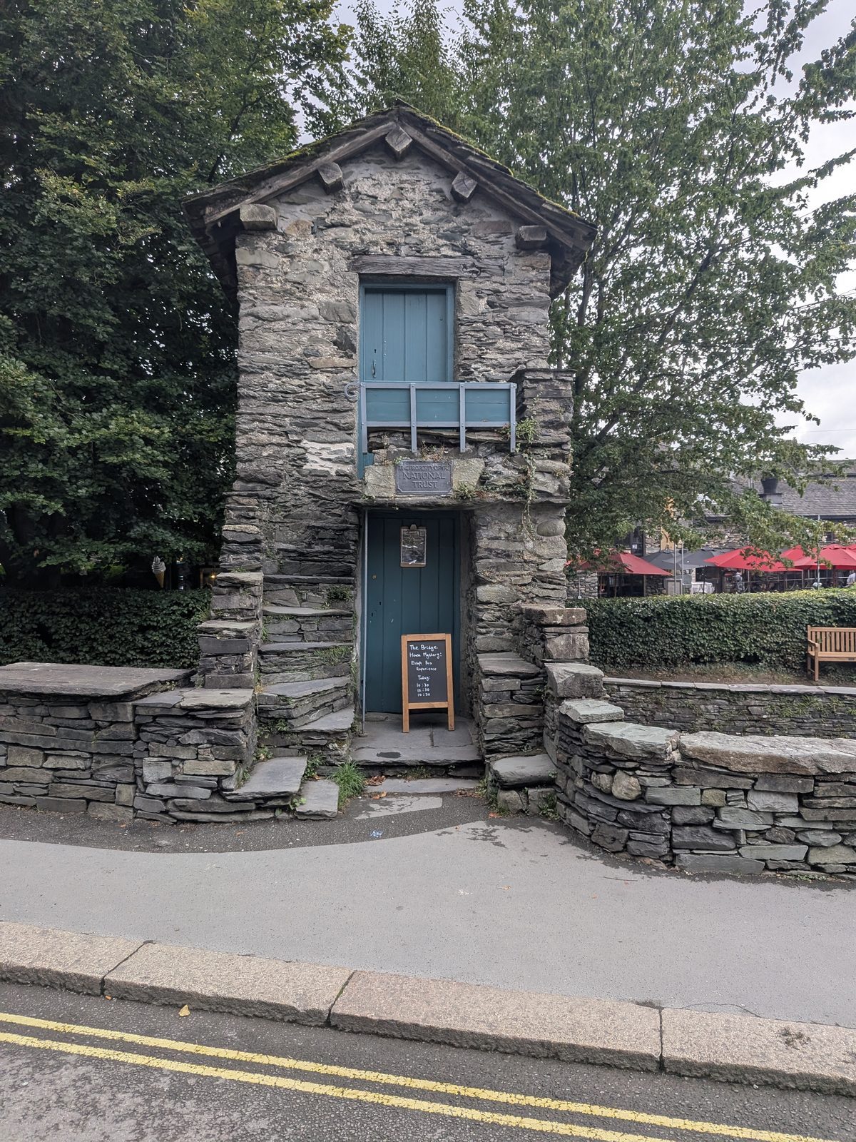 The front of Bridge House, Ambleside: stone steps climbing to a blue first-floor door, with the bridge arch below and trees behind