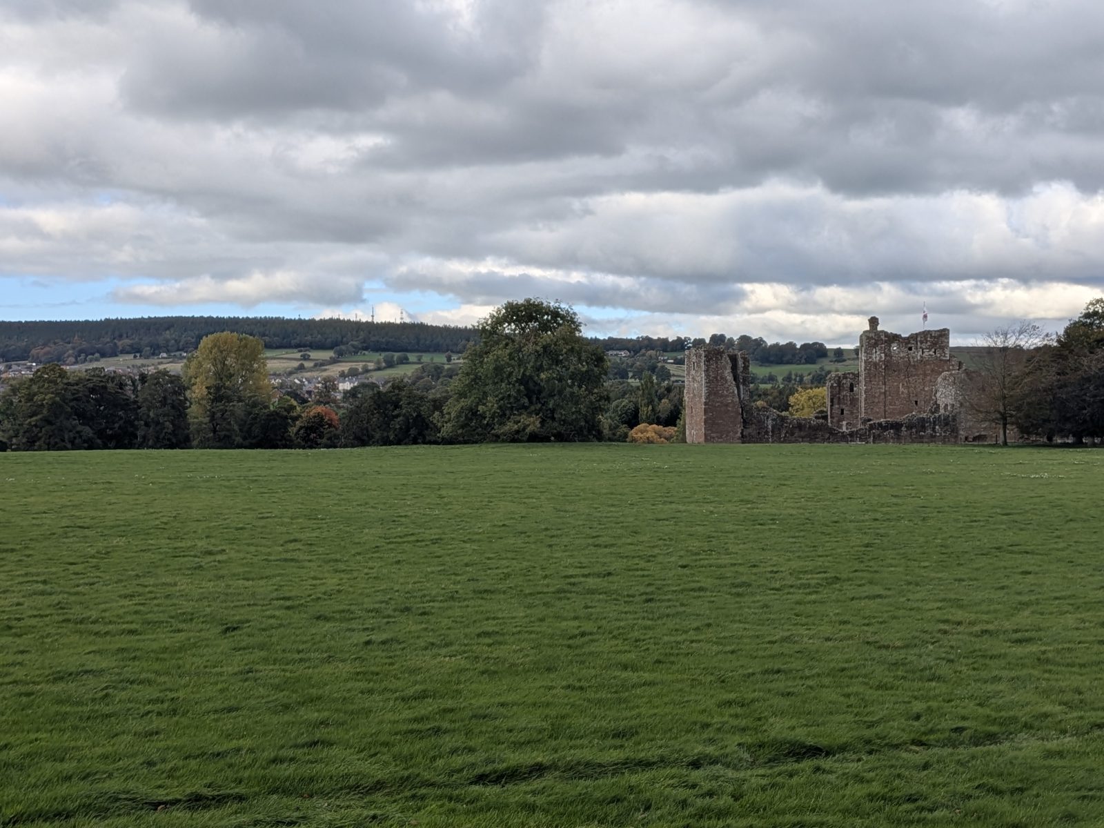 Brougham Castle on the skyline at the far side of a wide green field, with a town and wooded hill in the distance
