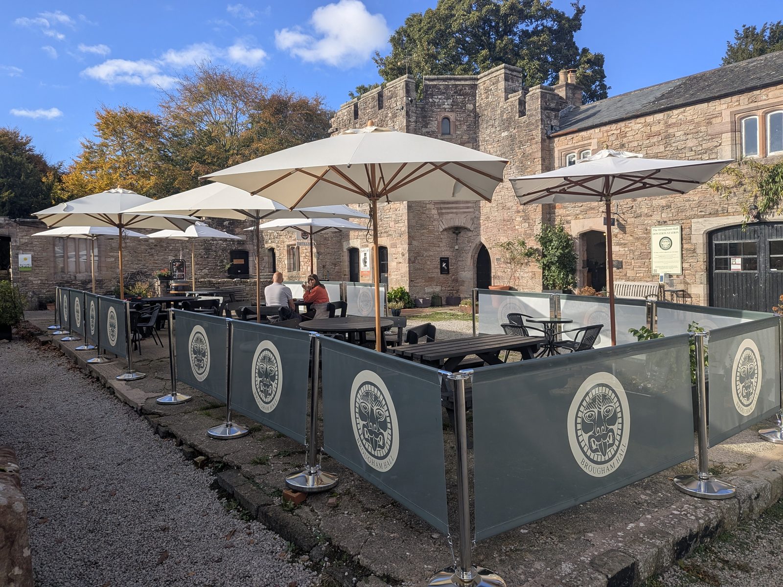 The Hideaway Café terrace in the Brougham Hall courtyard, with tables under cream umbrellas inside screens printed with the hall's emblem