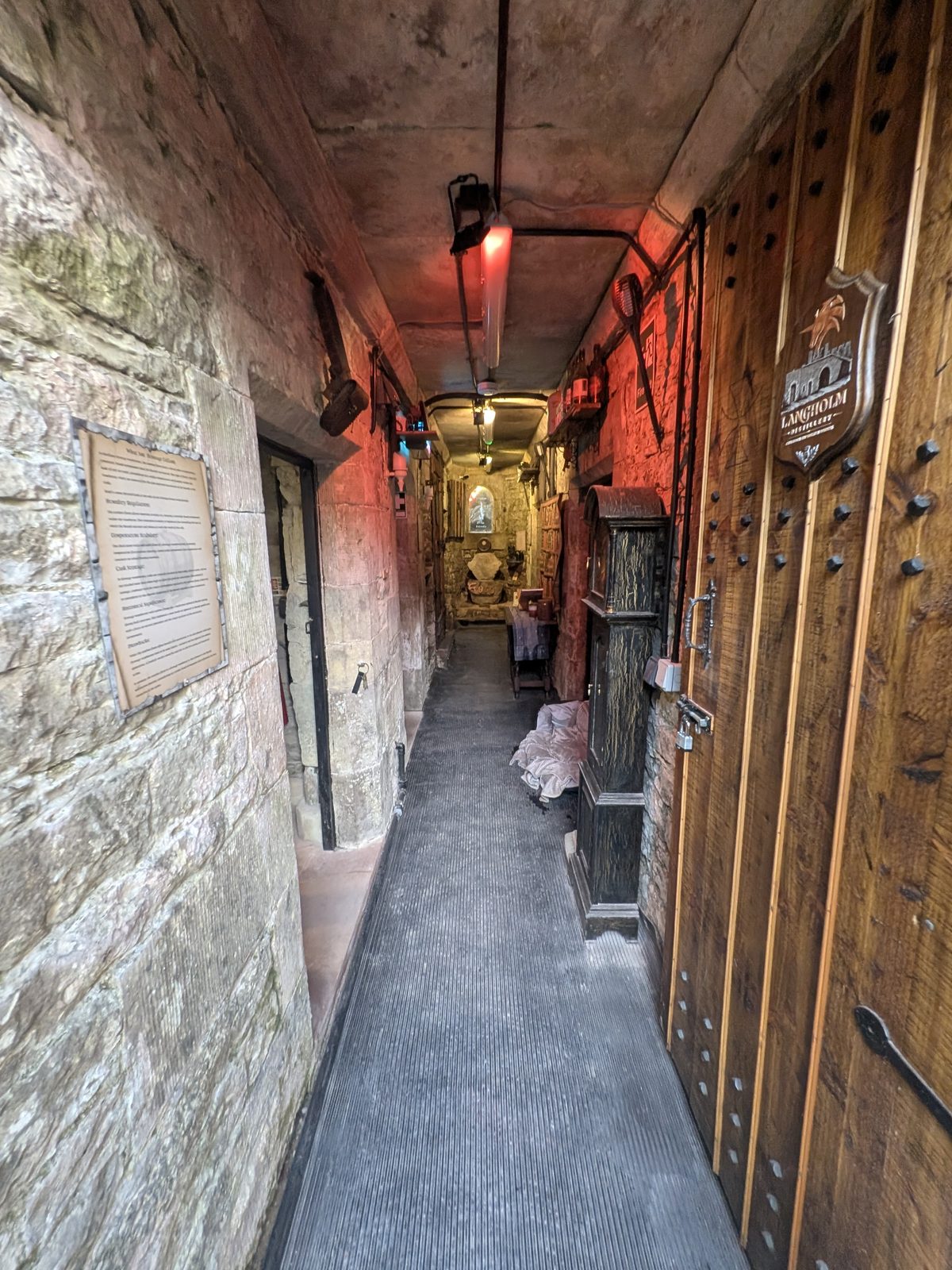 A long, narrow stone passage at Brougham Hall lit by red lamps, with a heavy studded door and the Langholm Distillery sign