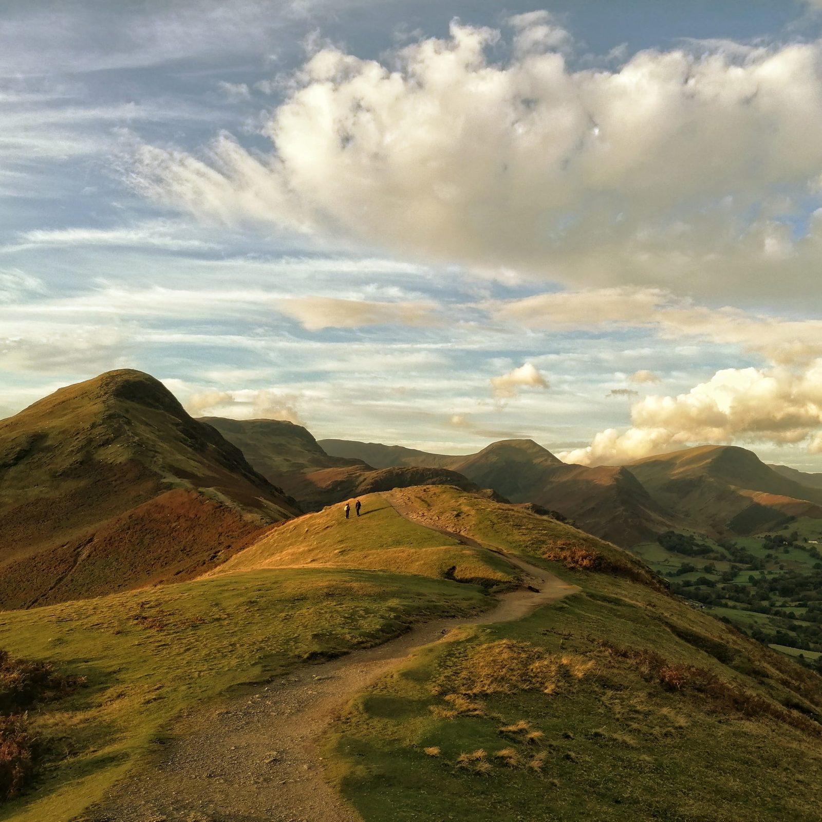 Two walkers on the grassy Catbells ridge path in low golden light, with fells and green valley beyond, Lake District