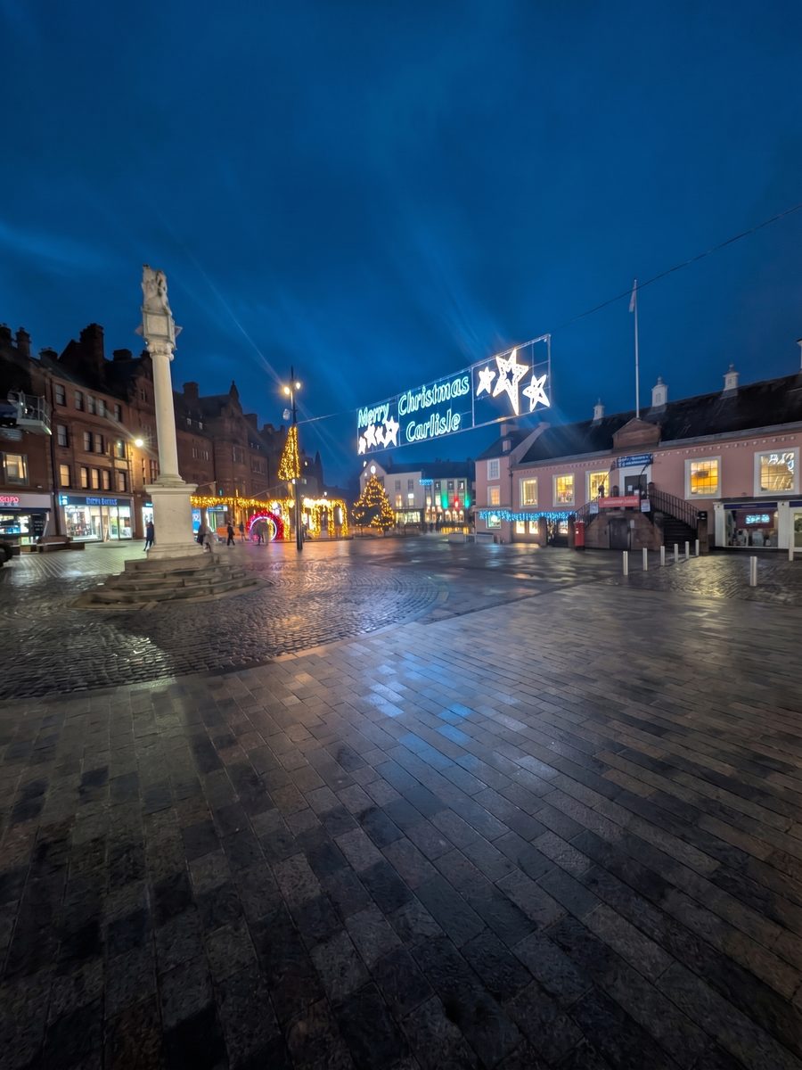 Carlisle's market square at dusk after rain, the wet paving reflecting a large illuminated Merry Christmas Carlisle sign with white stars above the shops, the market cross on the left and a lit Christmas tree beyond