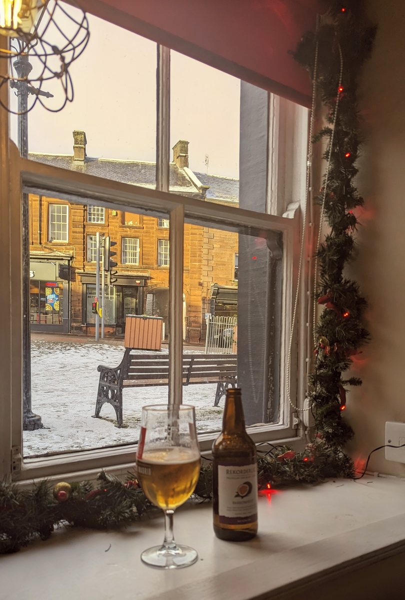 A pub window seat framed by a garland of fairy lights, a glass of beer on the sill, looking out over a snow-covered square with a bench and stone buildings in a Cumbrian market town