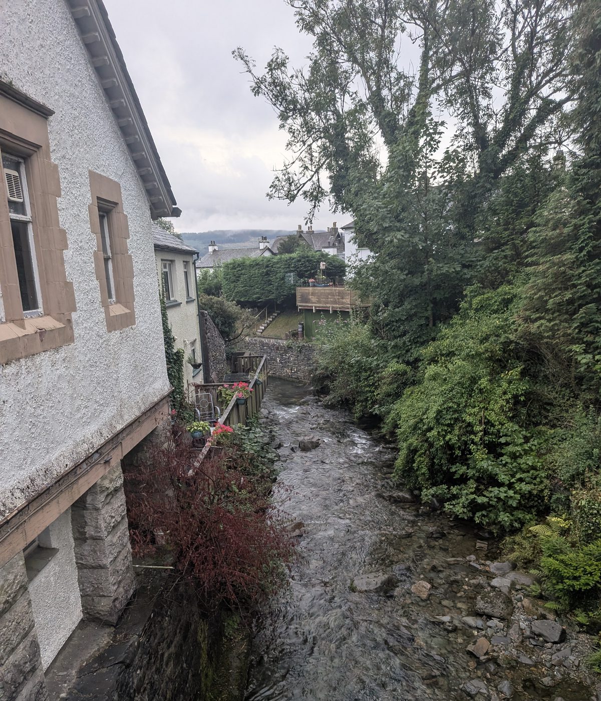 A beck running past the back of a stone building in Coniston, trees overhanging the water