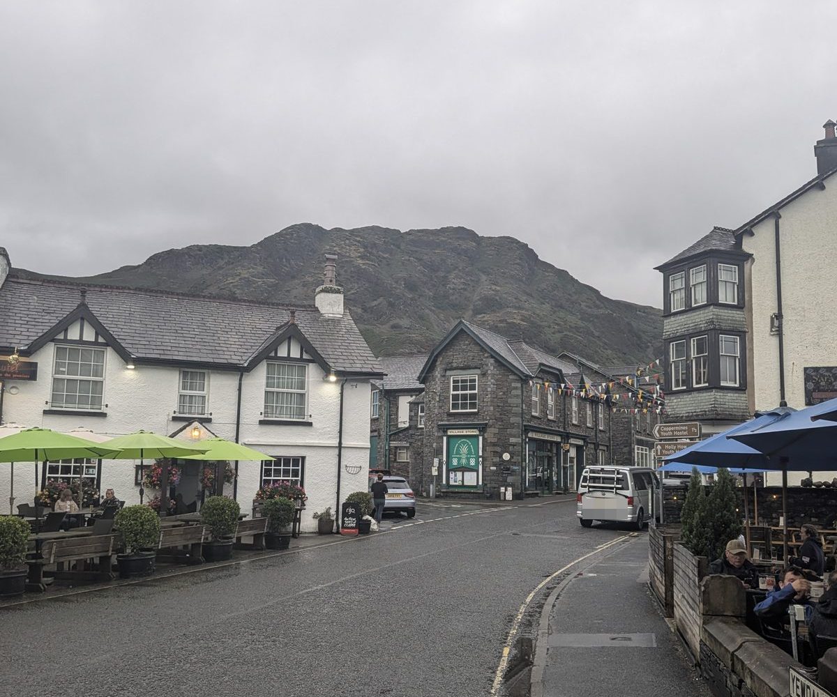 Coniston village centre: whitewashed and slate buildings with green parasols outside, a rocky fell rising straight behind the rooftops