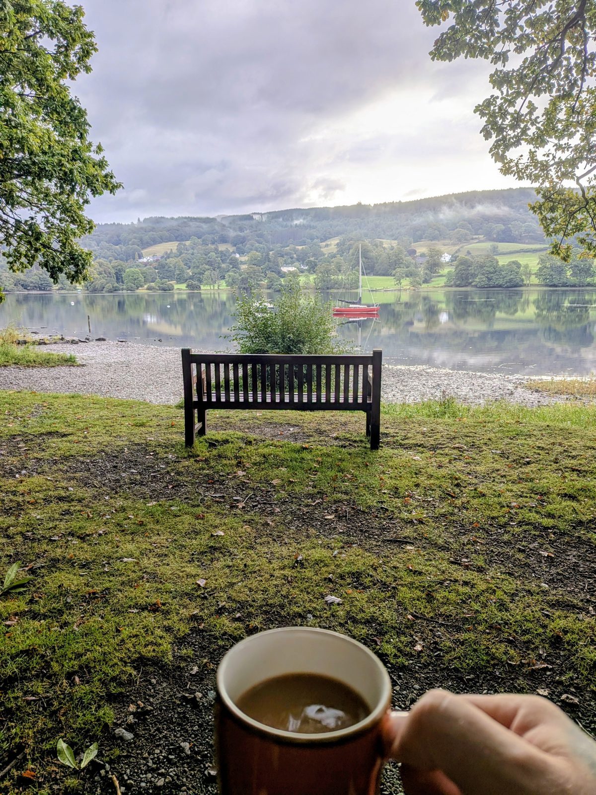 A hand holding a mug of coffee in front of a bench on the grassy shore of Coniston Water, the lake and wooded hills beyond
