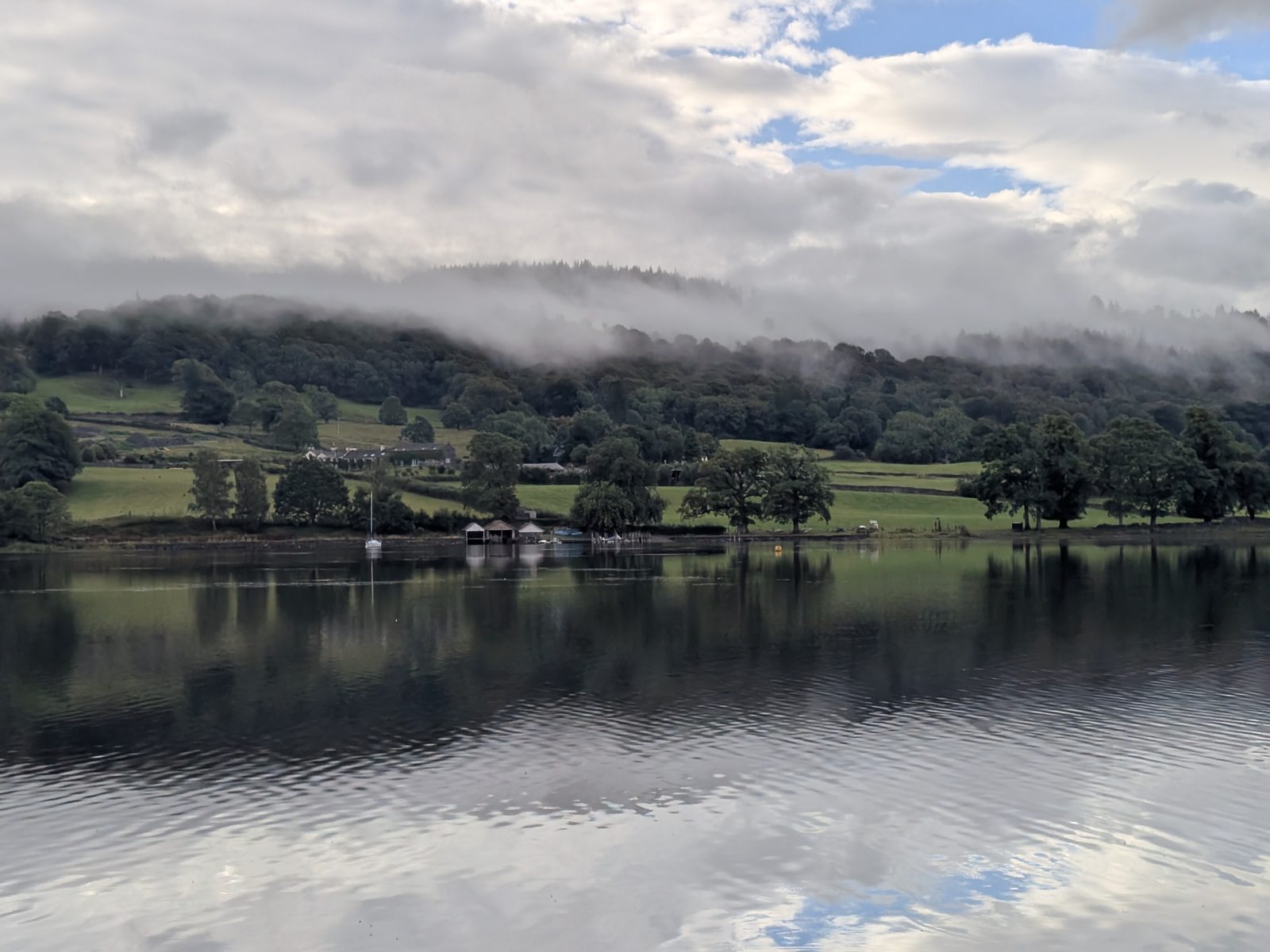 Coniston Water under low cloud, wooded shore and fields on the far side reflected in the lake