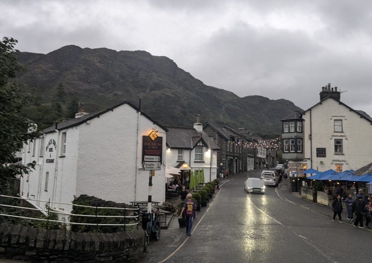 A street in Coniston with whitewashed buildings on the left, people on the pavement and a fell rising behind under grey cloud