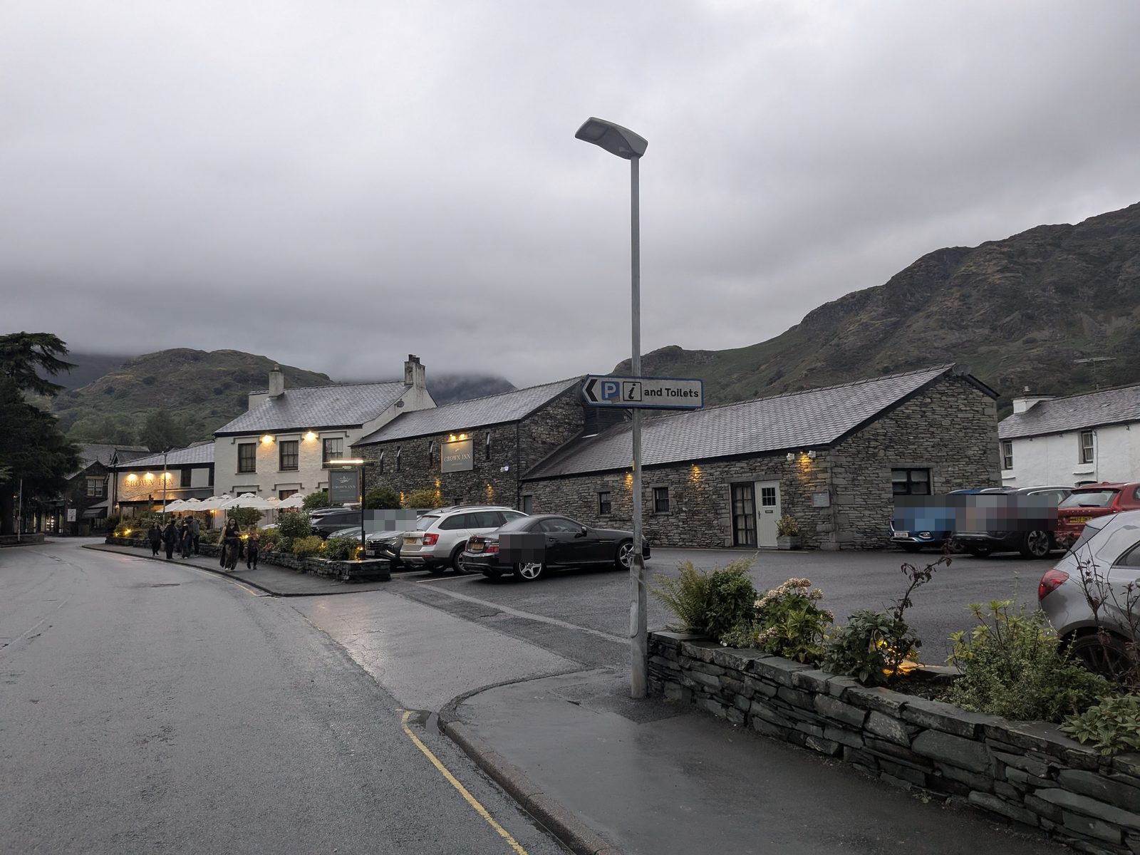 A wet road through Coniston at dusk, a slate pub with its lights on, cars parked outside and fells behind
