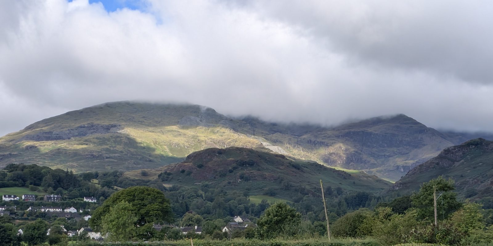 Rooftops and trees of Coniston village with the high fells rising behind, sunlight on the upper slopes