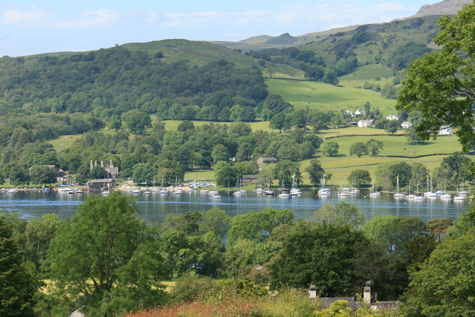 Yachts moored along the wooded shore of Coniston Water, with farmland and low fells rising behind