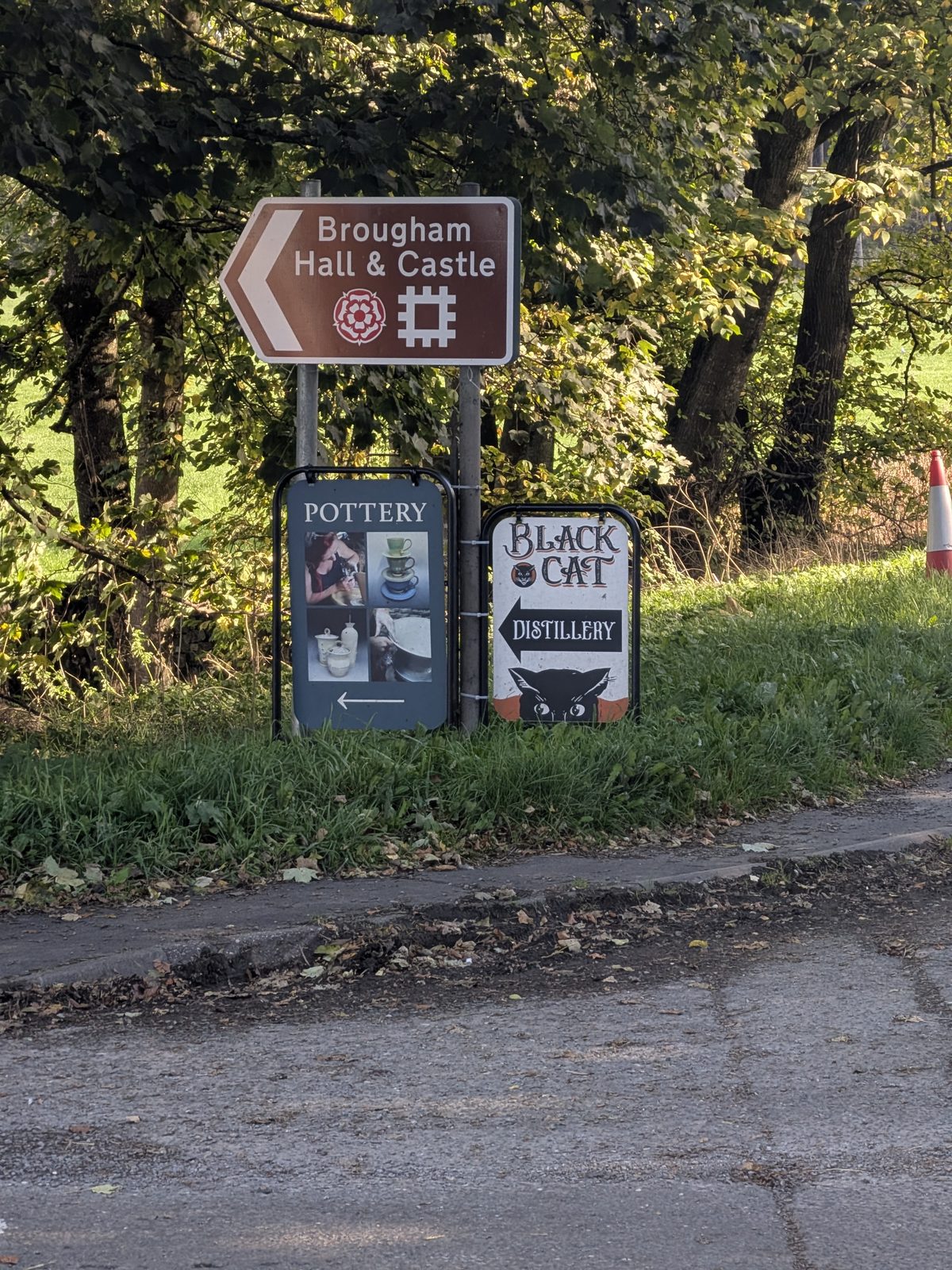 A brown road sign pointing left to Brougham Hall and Castle, above signs for the pottery and Black Cat Distillery