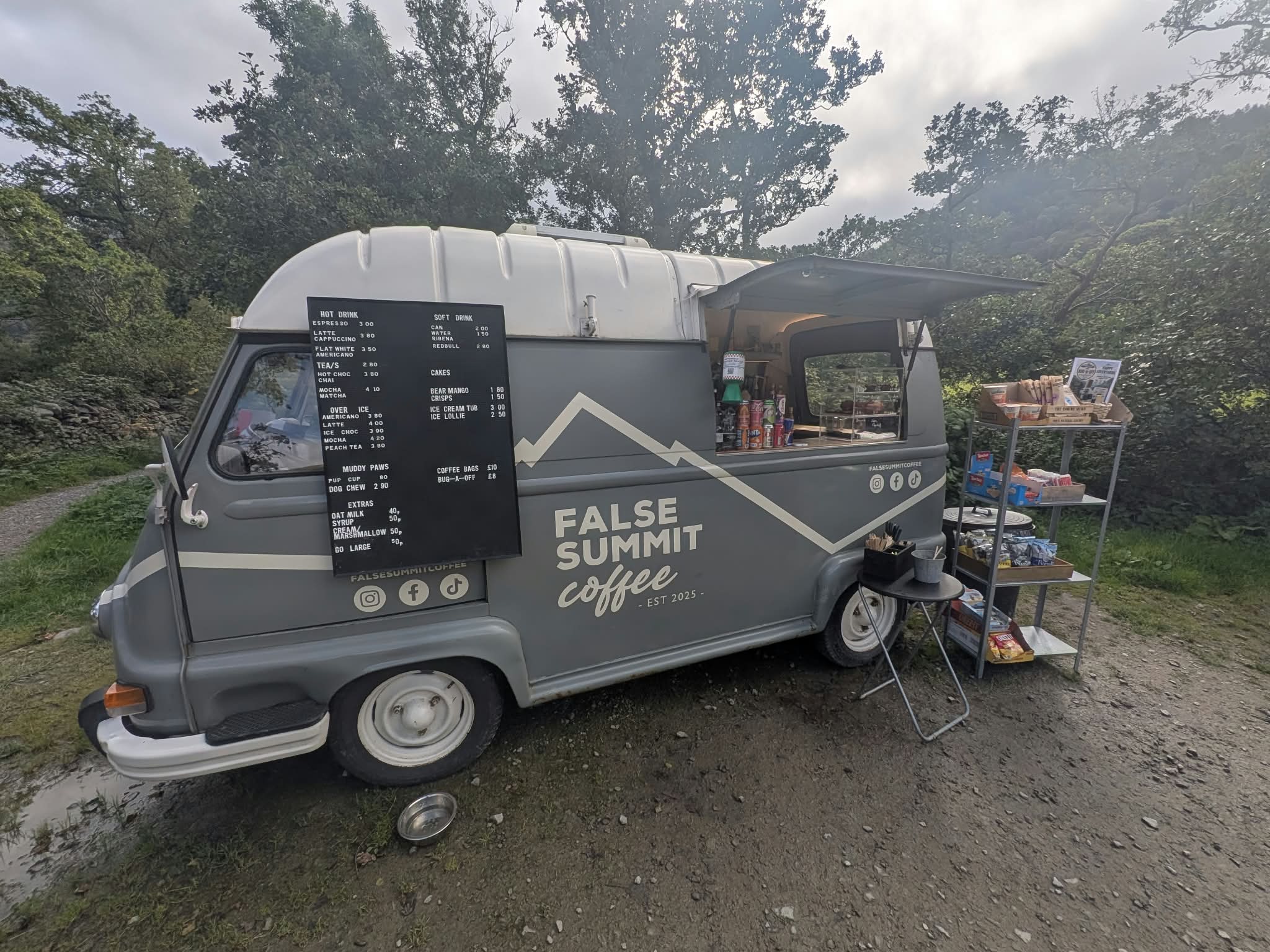 The grey False Summit Coffee van, a converted vintage van with its serving hatch open and a menu board, parked on gravel under trees at Glencoyne
