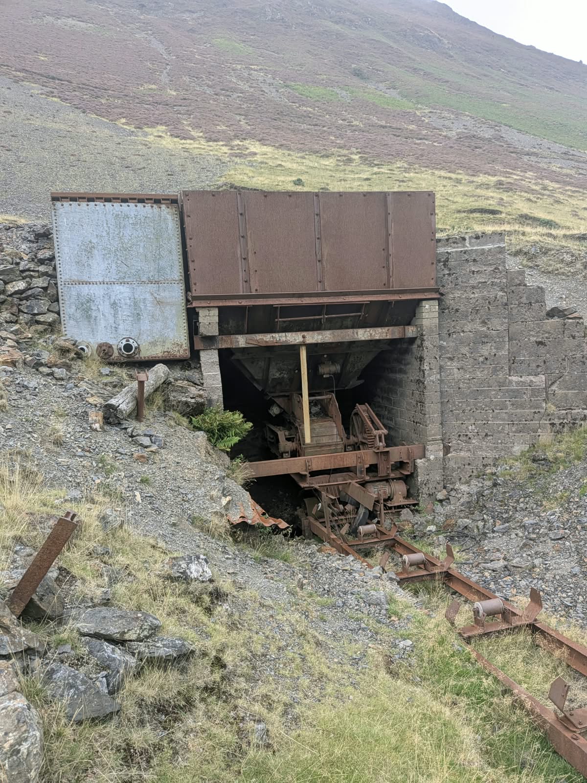 Rusted steel ore bin and machinery above a stone-lined opening, with rails in the foreground