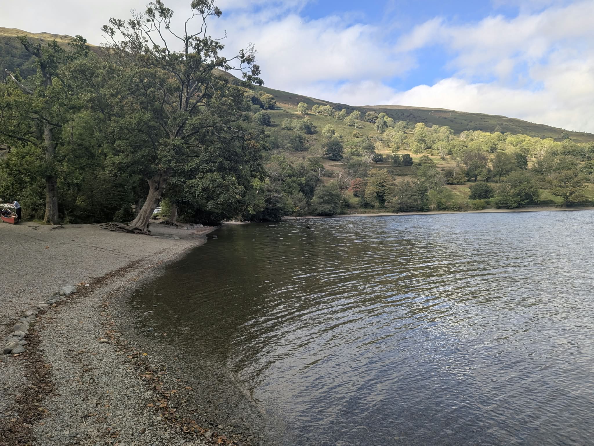 The shingle shore of Glencoyne Bay on Ullswater, trees leaning over the water and a canoe pulled up on the beach, with the wooded fellside beyond