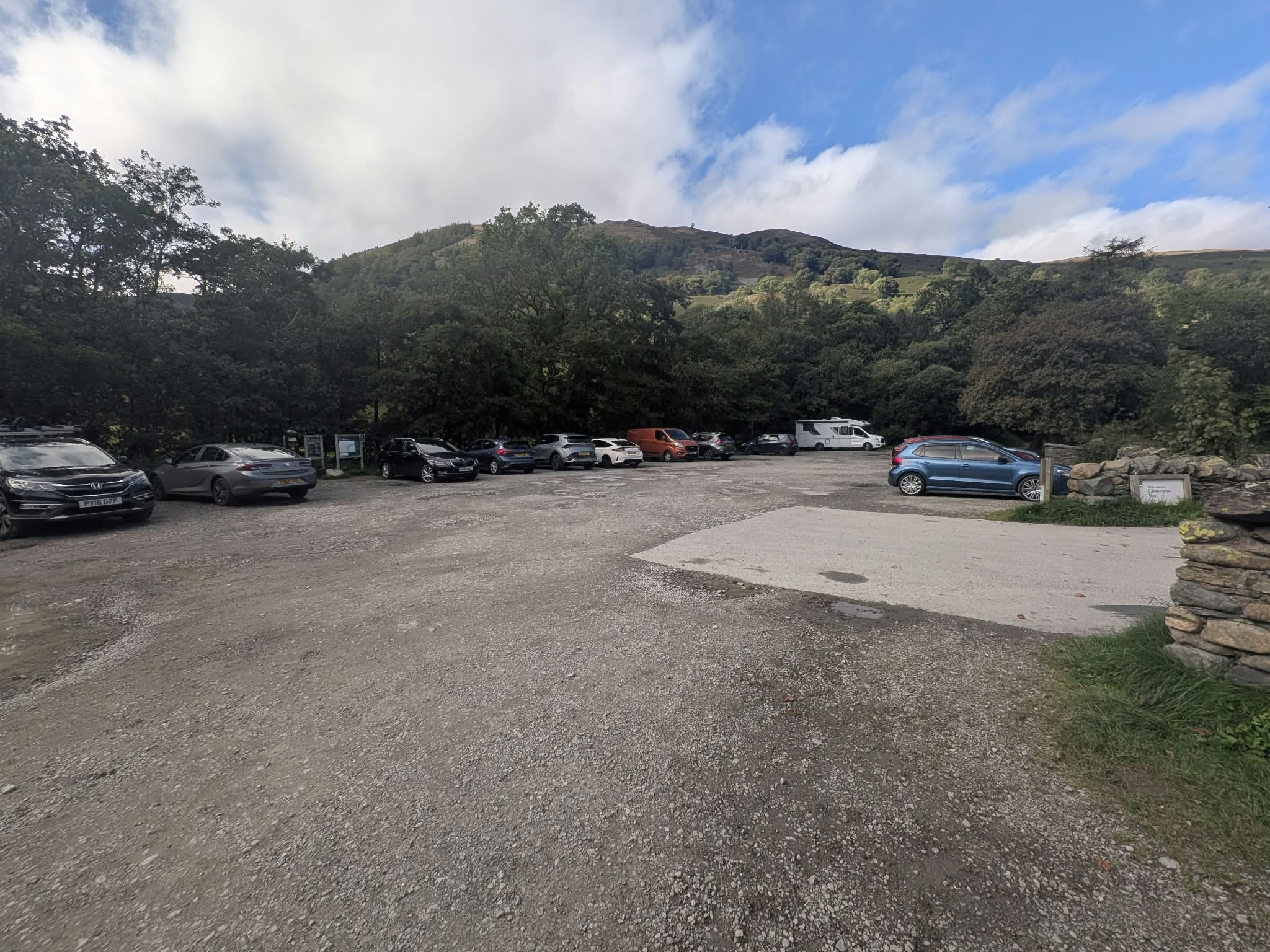 The gravel National Trust car park at Glencoyne, with cars parked along the trees and the fell behind
