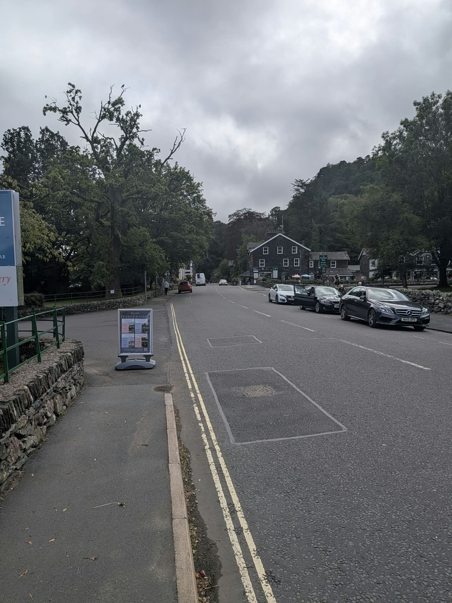 The main road through Glenridding, with a pavement sign and a line of cars parked along the far side under the trees