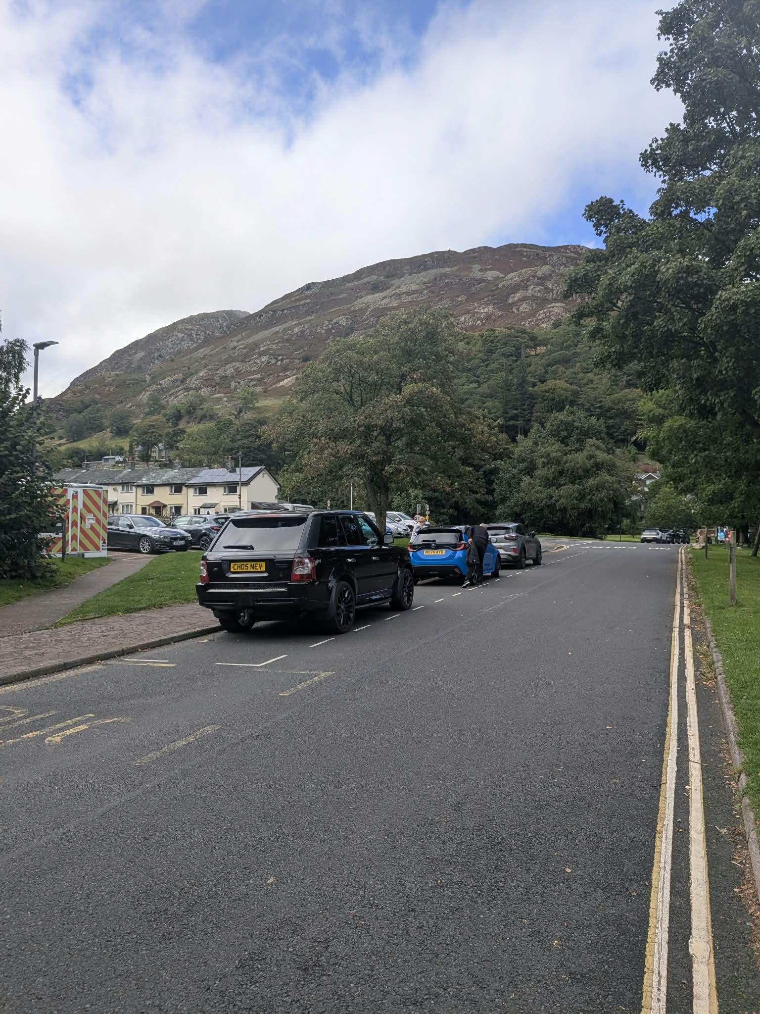 Cars parked along the road into Glenridding, with houses and a wooded fell ahead