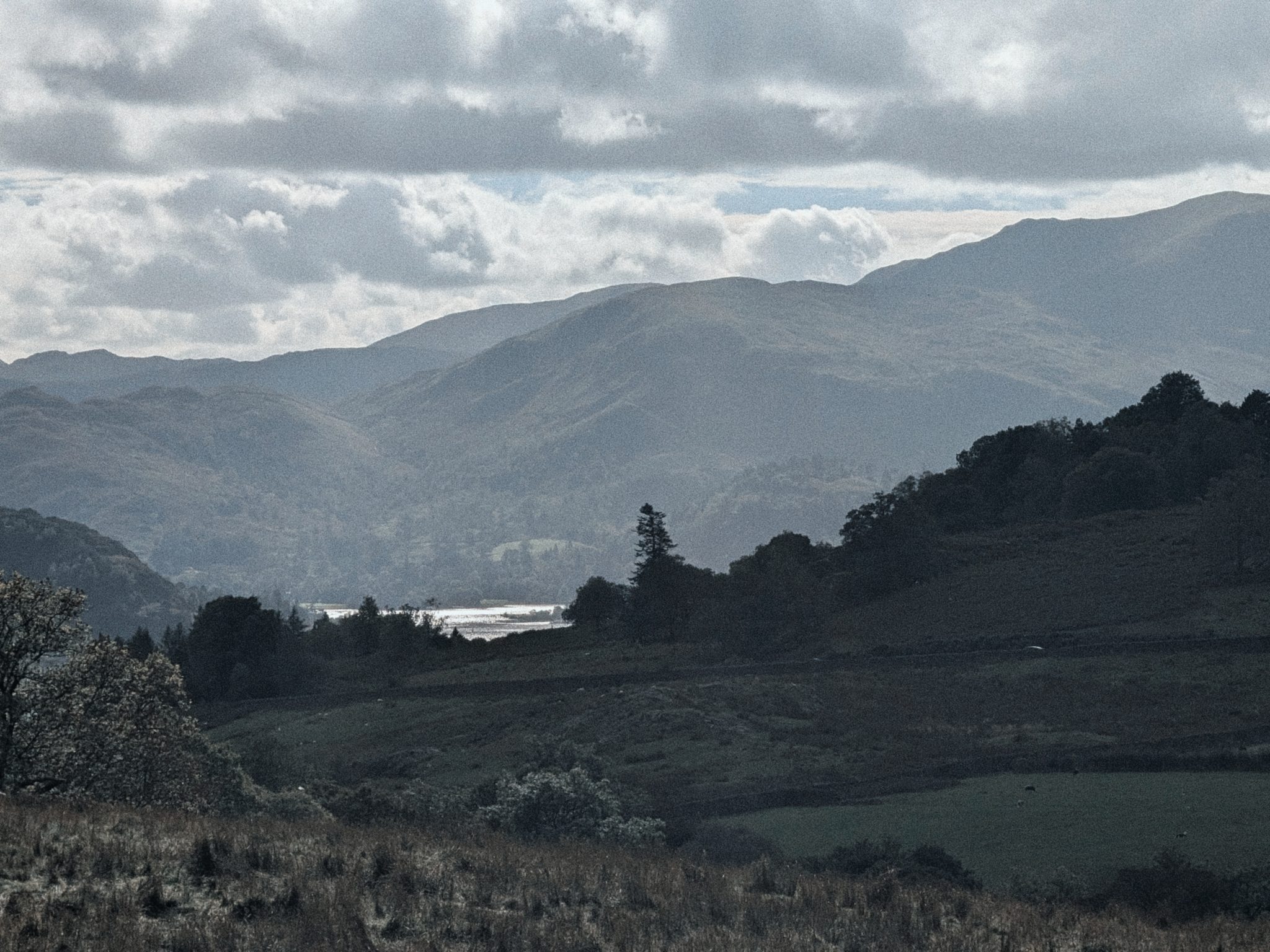 A distant strip of lake glinting between dark wooded hills under heavy cloud, with layers of fells beyond.