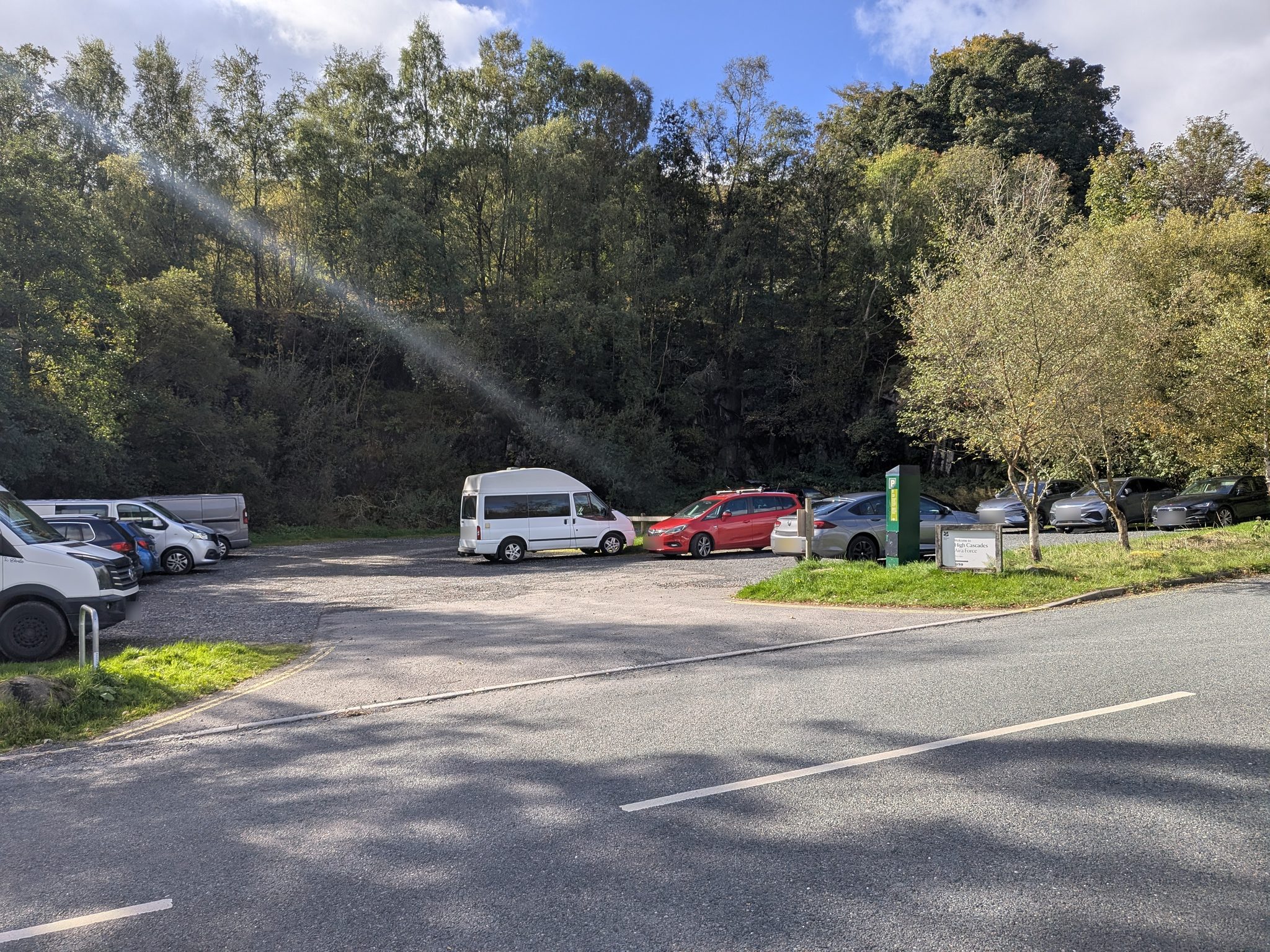 A small tree-lined car park seen from the road, with parked cars and a pay station.