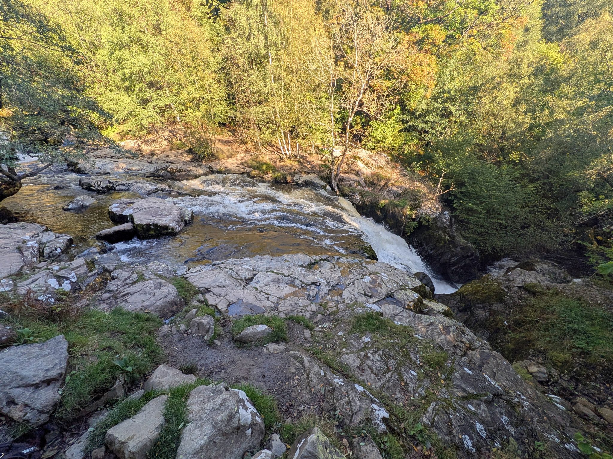 Water sliding over pale rock slabs and dropping out of sight, with autumn birch trees on the far bank.