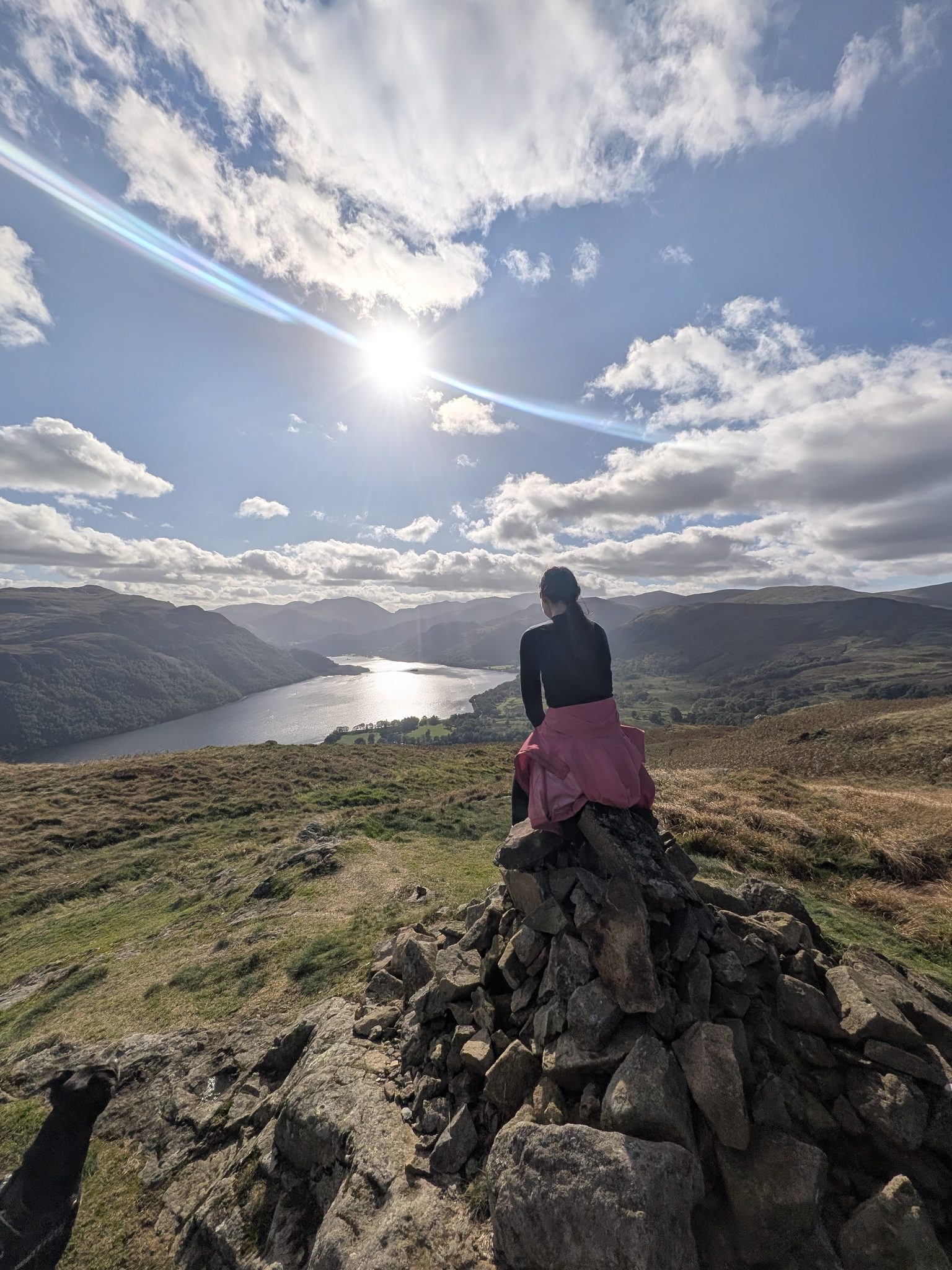 A woman seen from behind sitting on a stone cairn, looking down a long lake towards distant fells with the sun overhead.