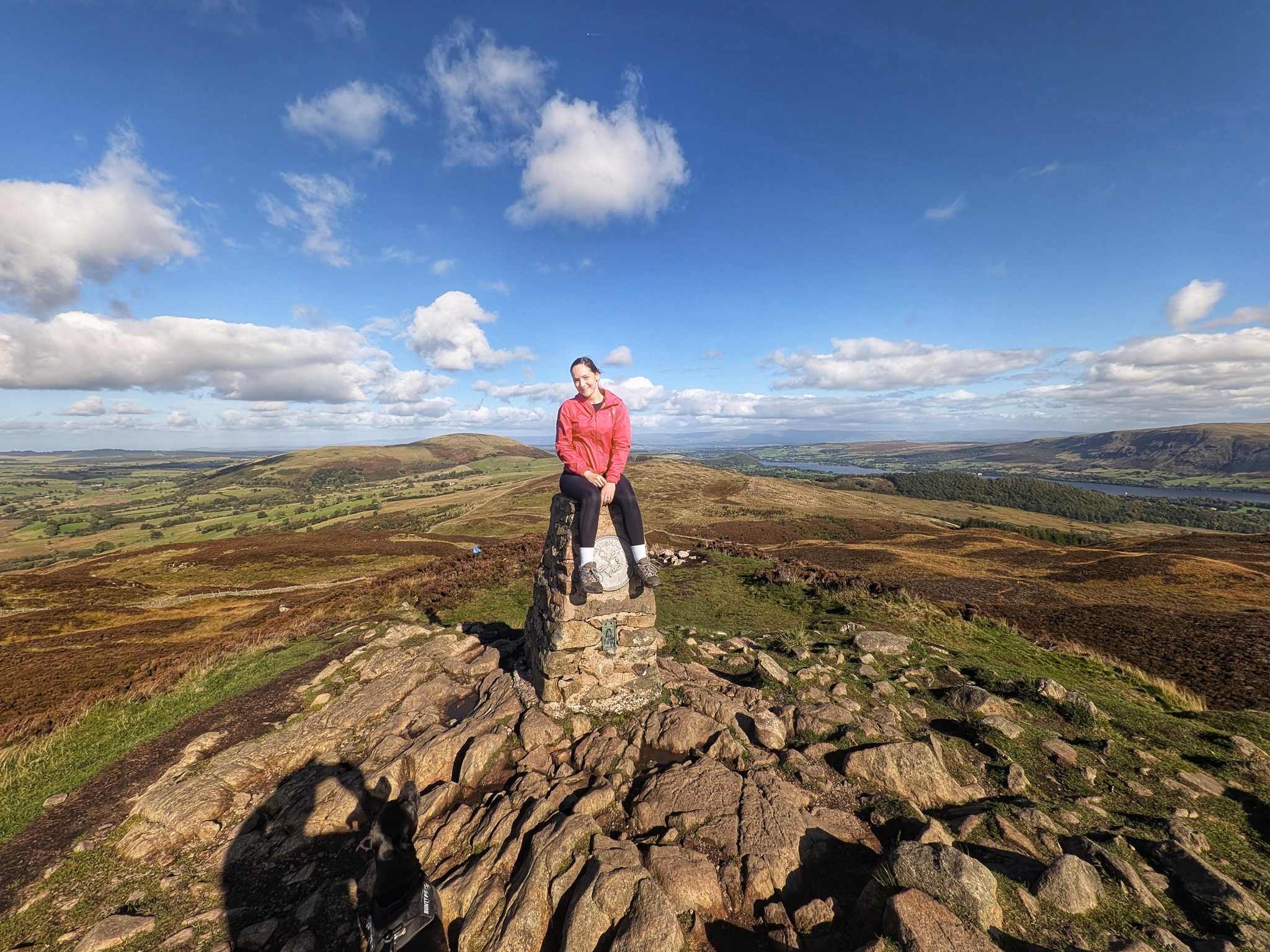 A woman in a red jacket sitting on a stone trig pillar on a rocky summit under a blue sky.