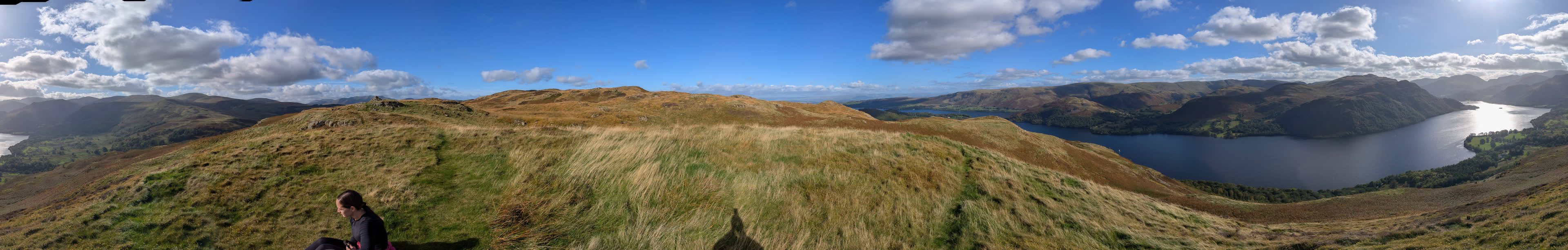 A wide panorama across grassy fell top to a long lake curving between fells.