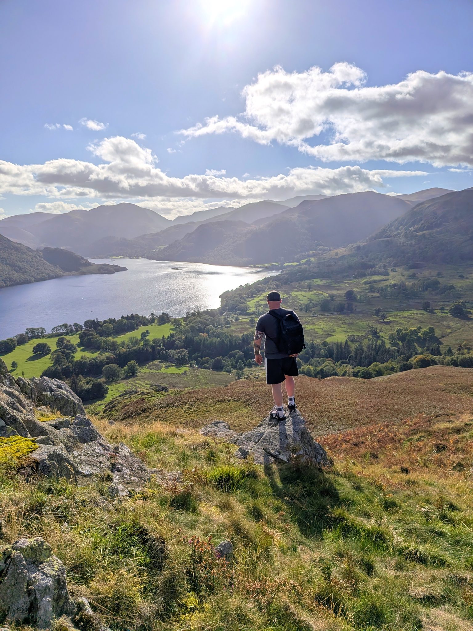 A man with a rucksack seen from behind, standing on a rock above green fields and the head of a long lake.