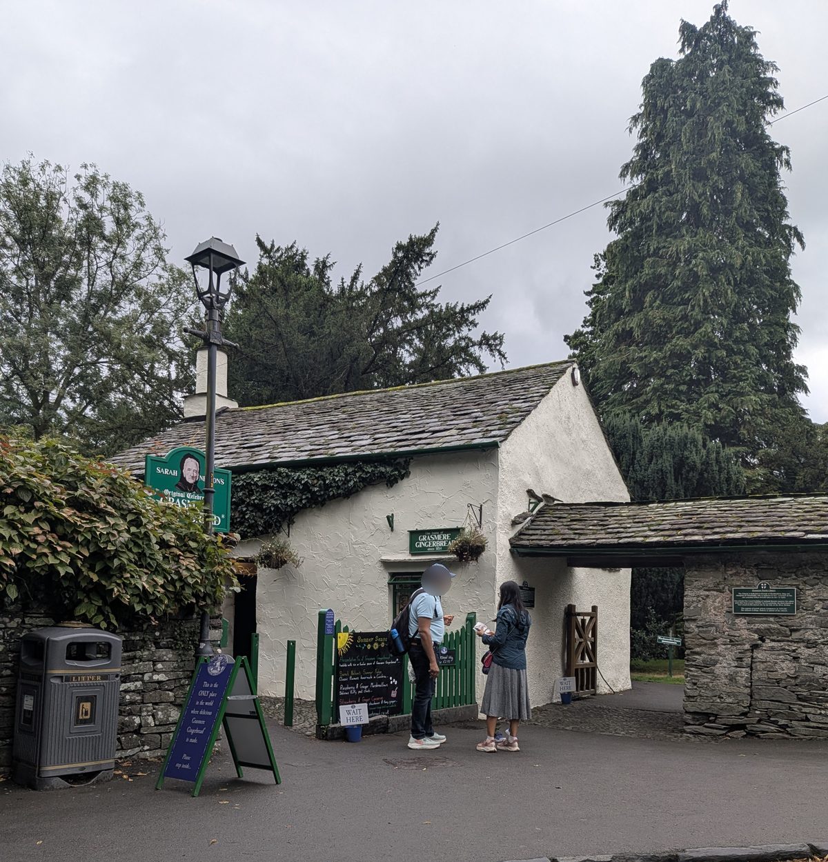 Sarah Nelson's Grasmere Gingerbread shop: a whitewashed cottage with a slate roof and green railings, people at the door, and the lychgate into the churchyard beside it