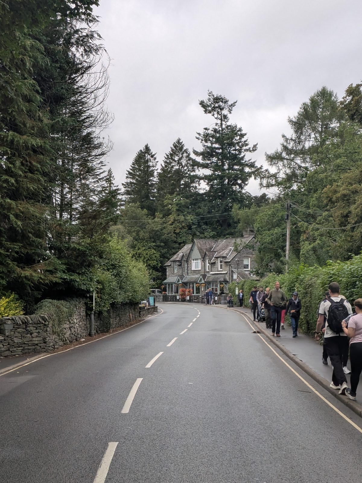 The approach road into Grasmere village, walkers on the pavement and a slate building among trees ahead