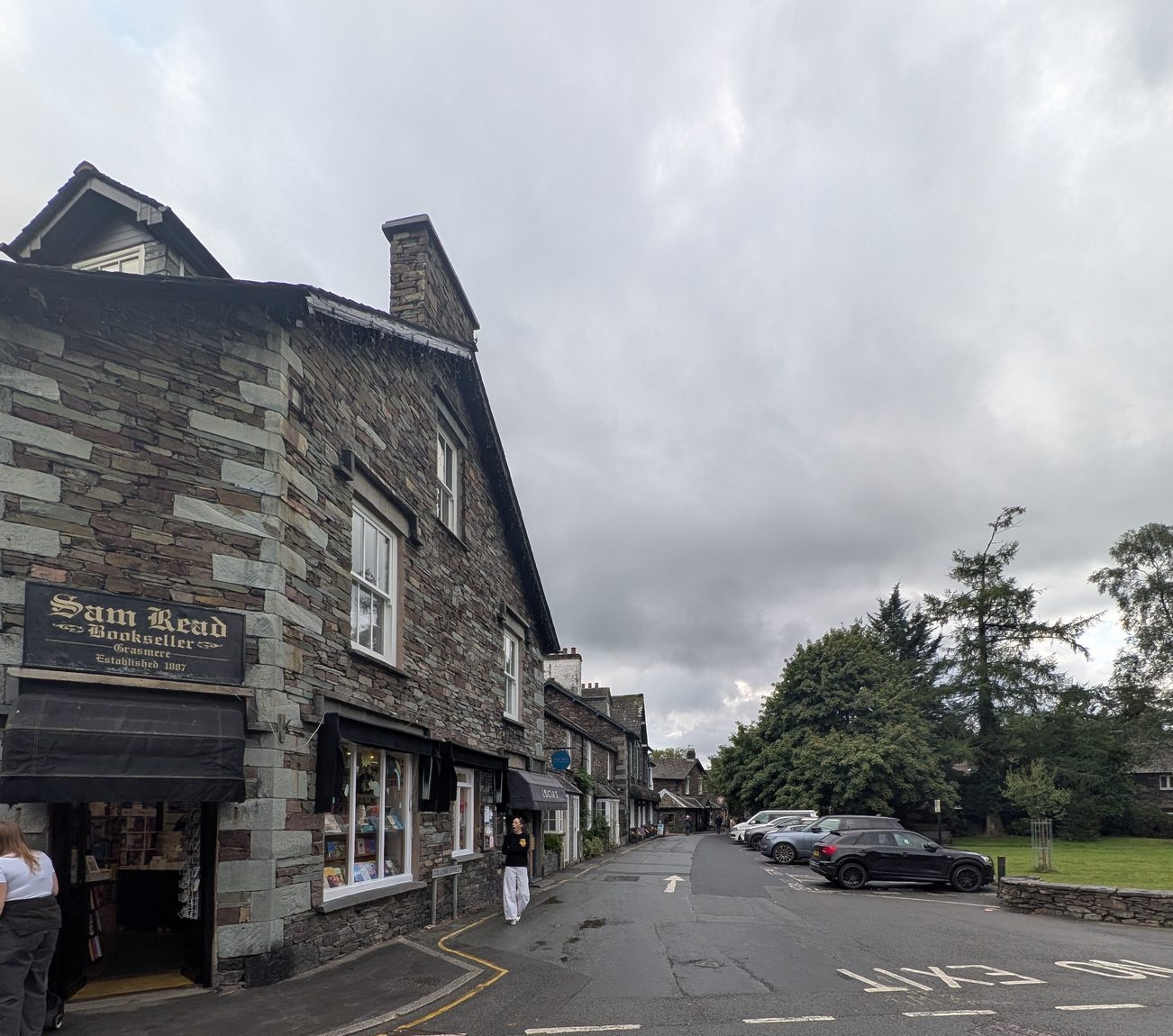 Grasmere village centre: slate buildings with shop awnings along the street, parked cars and trees beyond