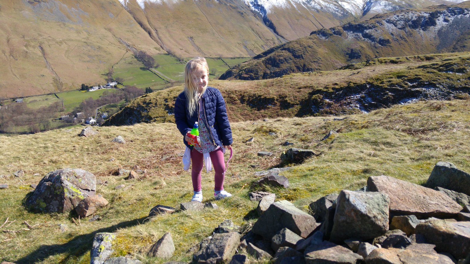 A young child walking the easy grassy slopes of Hallin Fell, the Martindale valley and snow-topped fells behind