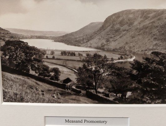 Old sepia photograph of the original Haweswater lake with the Measand promontory reaching into the water, walled fields and trees in the foreground and the fells rising behind, captioned 'Measand Promontory'