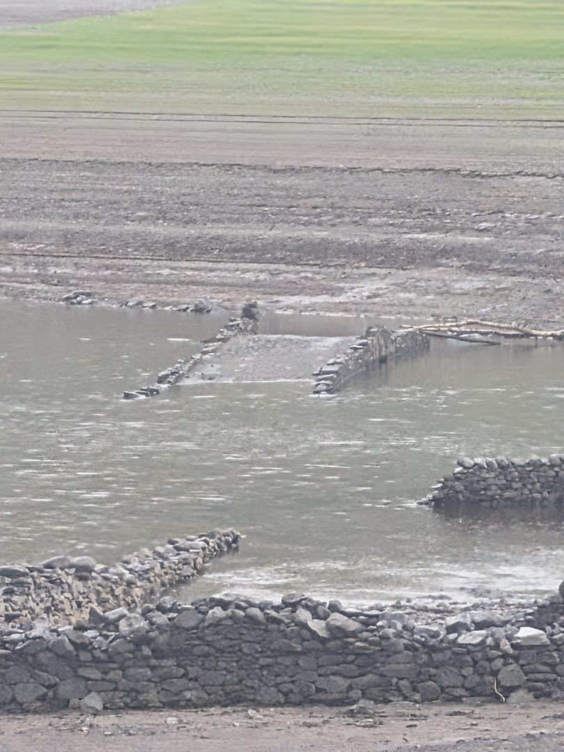 The tops of two stone bridge parapets just breaking the surface of grey water on the bed of Haweswater, with drystone field walls emerging from the water in the foreground and bare silt beyond