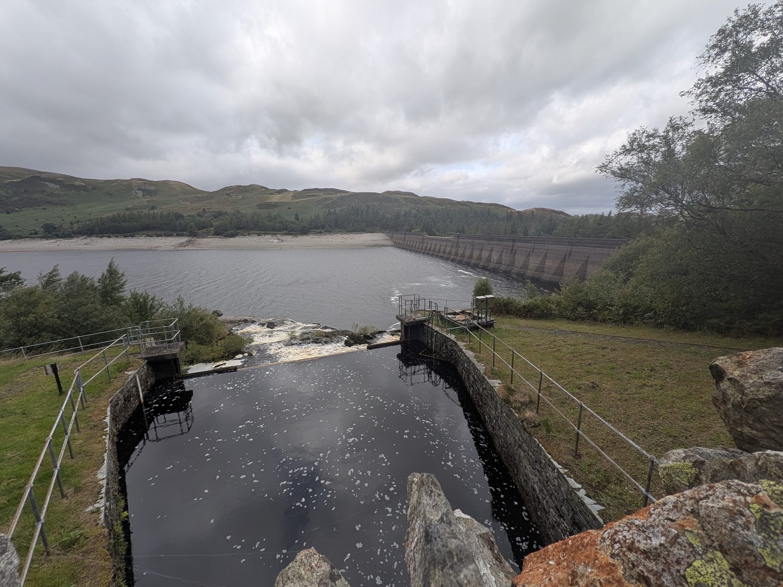 Looking along the top of the Haweswater dam from the viewing area, with the concrete spillway channel and railings in the foreground, the long straight line of the dam wall stretching away and the reservoir beyond under a grey sky