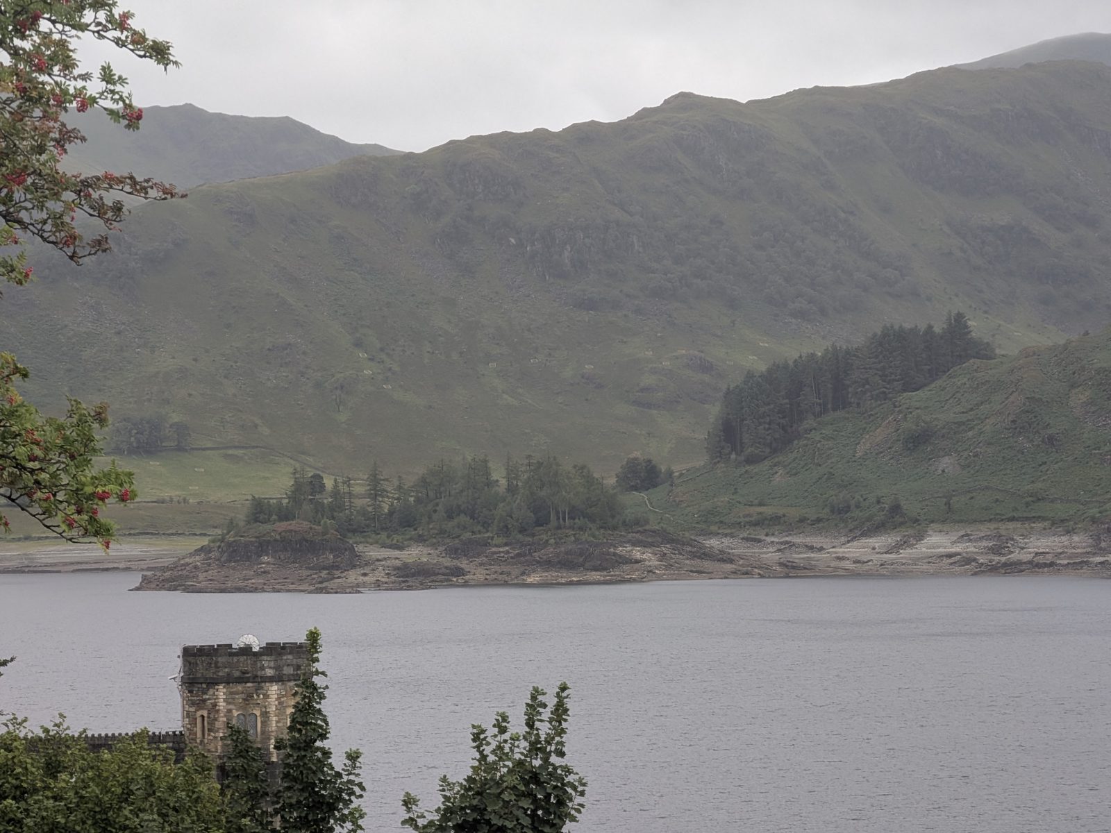 The castellated stone draw-off tower at the water's edge of Haweswater in the foreground, with the grey reservoir behind it, the wooded island of Wood Howe and the exposed pale shoreline beyond, and the fellside rising above