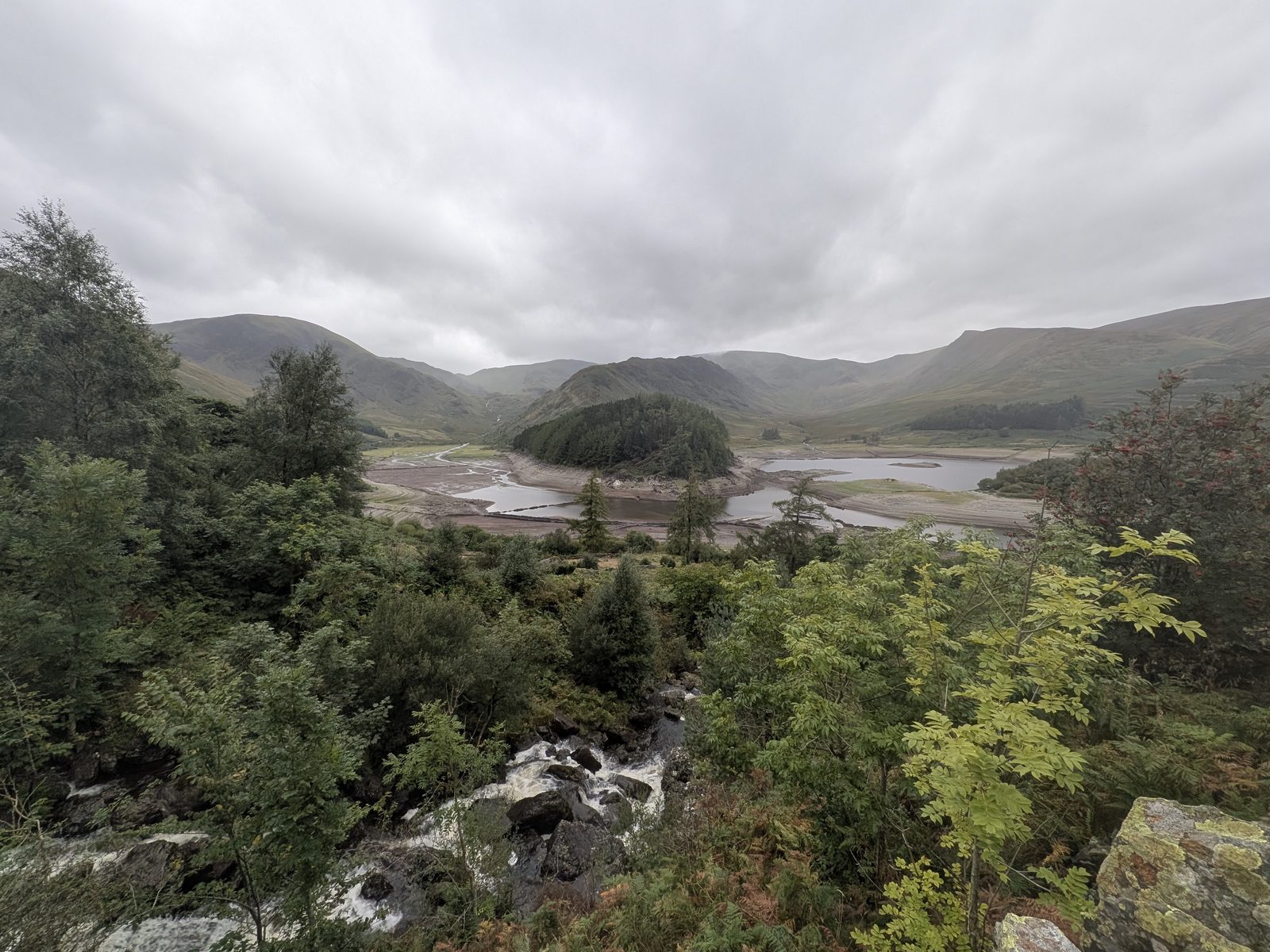 Haweswater from above the head of the reservoir under a grey sky: the wooded island of Wood Howe in the centre with the pale drawn-down bed exposed either side of it, a beck tumbling over rocks in the foreground, and the fells of Riggindale and High Street behind