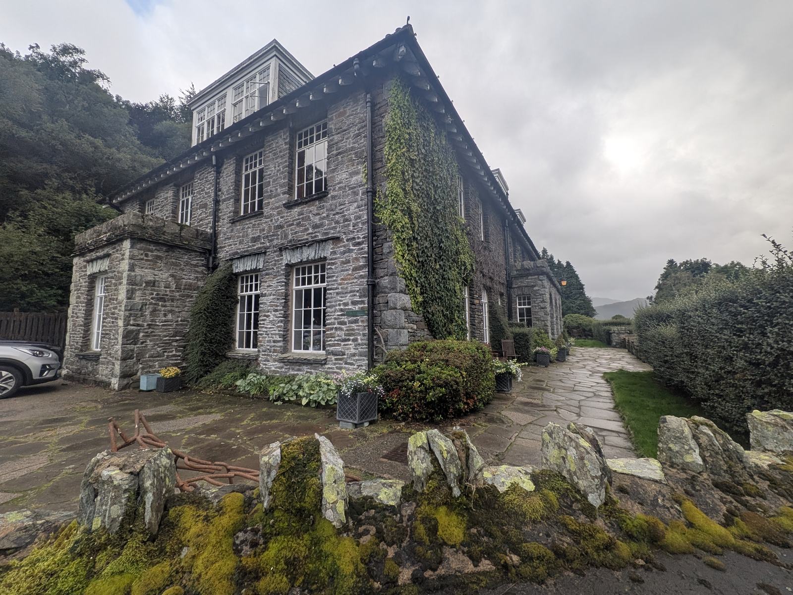 The Haweswater Hotel from outside: a long two-storey stone building with rows of white-framed windows, a slate roof, creeper on the walls, a flagged terrace and a low stone wall in front, under a grey sky