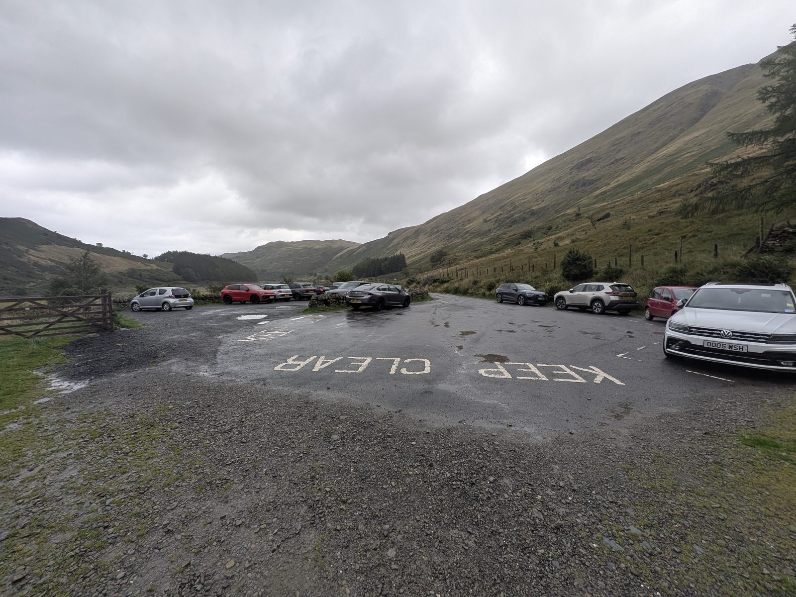Mardale Head car park at the end of the Haweswater road: a rough surfaced area with KEEP CLEAR painted on the turning space, cars parked around the edge, a wooden gate on the left and the fellside rising steeply on the right under grey cloud