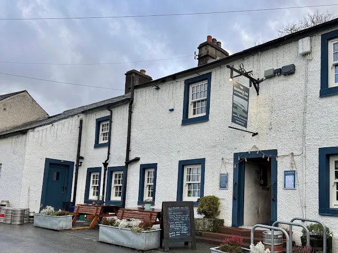 The Mardale Inn at Bampton: a long whitewashed pub with blue-painted window frames and doors, picnic benches and planters outside, a chalkboard by the door, under a grey sky