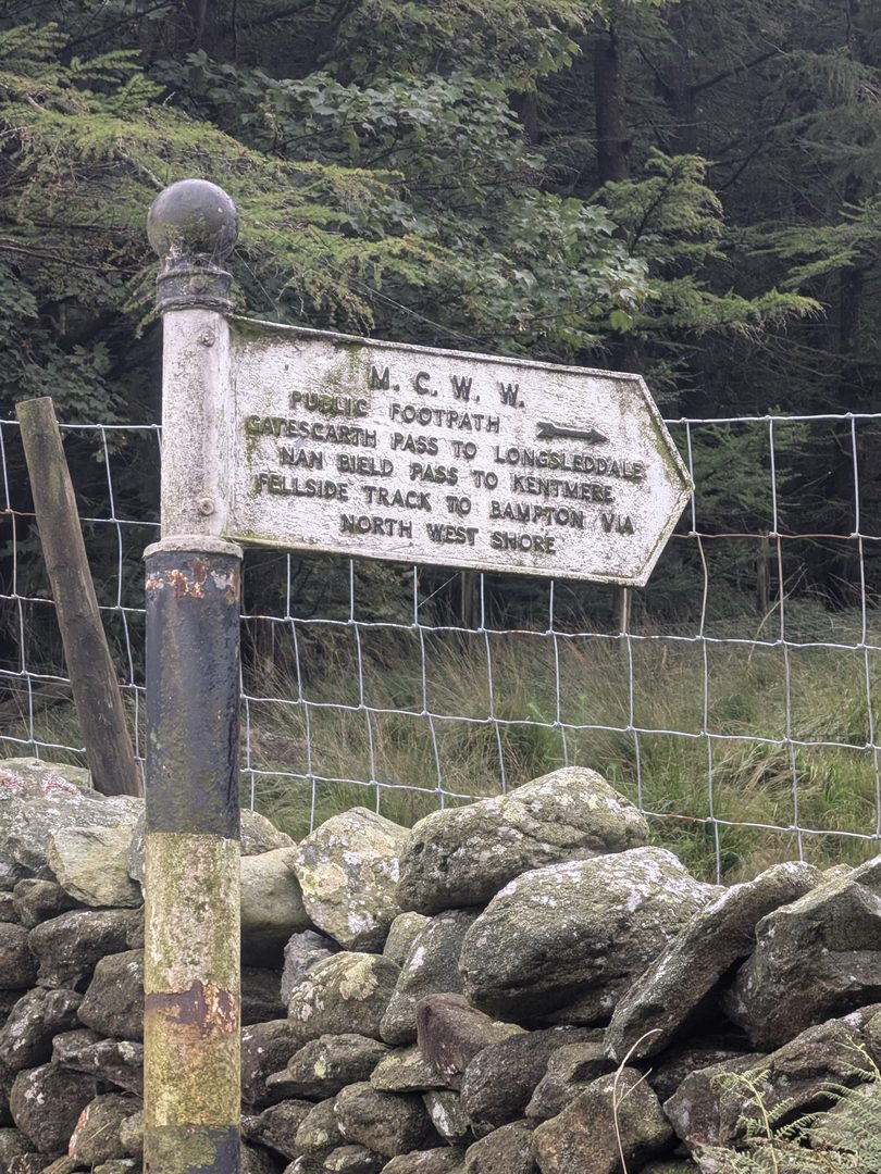 A white cast-iron M.C.W.W. fingerpost on a black post beside a drystone wall and wire fence at Haweswater, its arm lettered 'Public footpath — Gatescarth Pass to Longsleddale, Nan Bield Pass to Kentmere, fellside track to Bampton via north west shore'