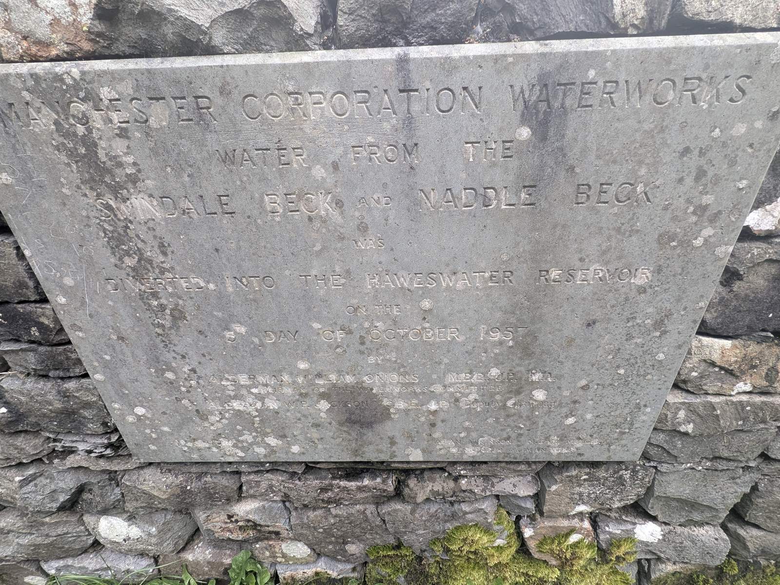 A weathered stone plaque set into a drystone wall, lettered in capitals: Manchester Corporation Waterworks — water from the Swindale Beck and Naddle Beck was diverted into the Haweswater Reservoir on the day of October 1955, followed by the names of the aldermen