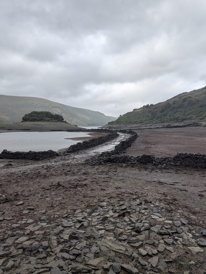 The old walled road of Mardale Green running out across the muddy bed of Haweswater towards the water, with the wooded island of Wood Howe beyond, and a slate-strewn bank in the foreground