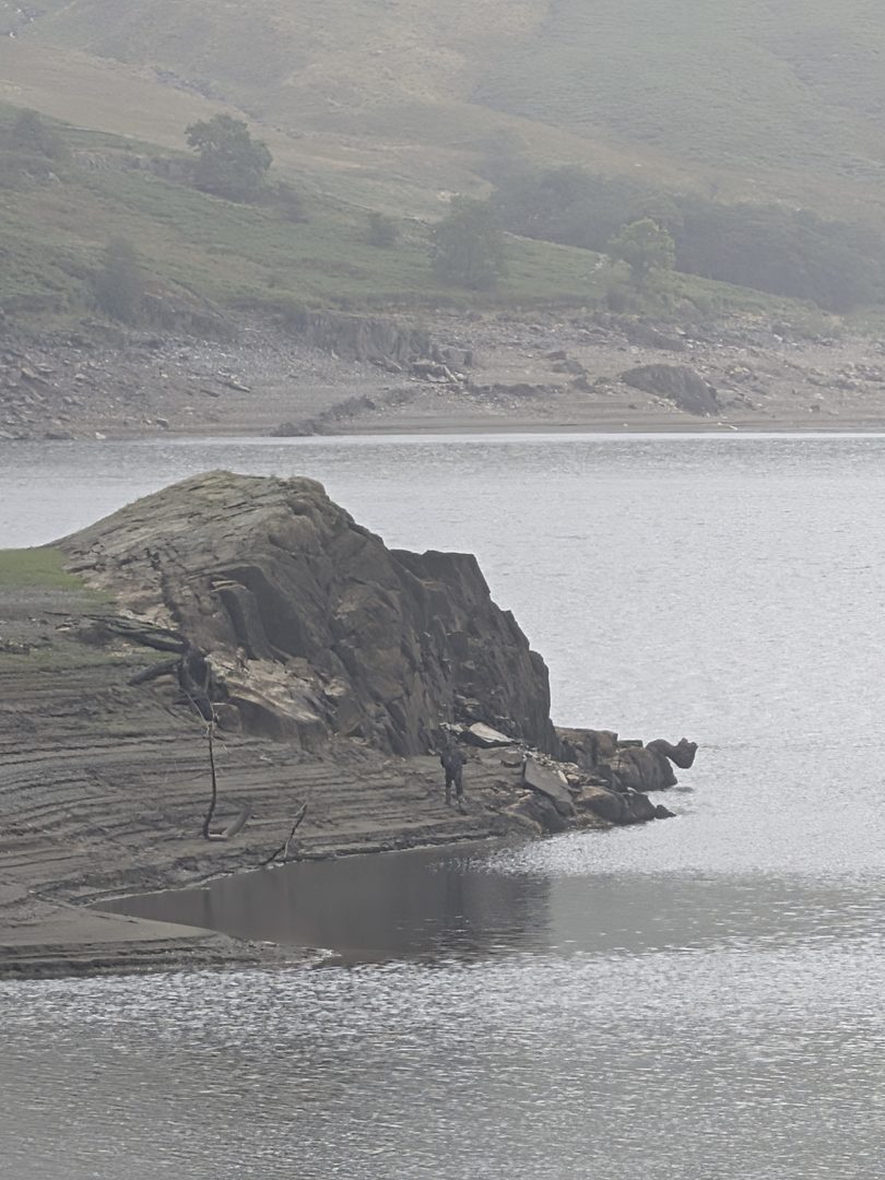 A bare, dark rock outcrop running out into the grey water from the exposed shore of Haweswater, with the pale drawn-down shoreline and the fellside beyond under low cloud