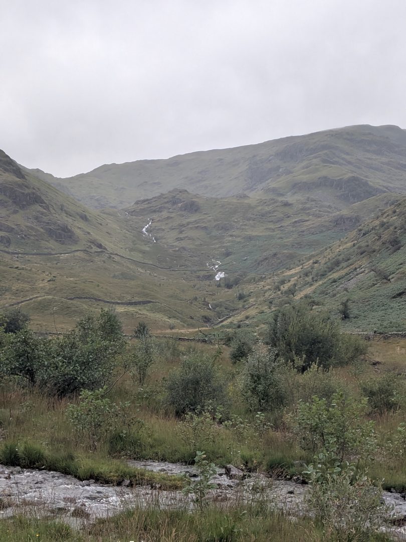 A beck coming down off the fellside at the head of Mardale in a series of small white falls between bracken and trees, seen from the exposed bed of Haweswater